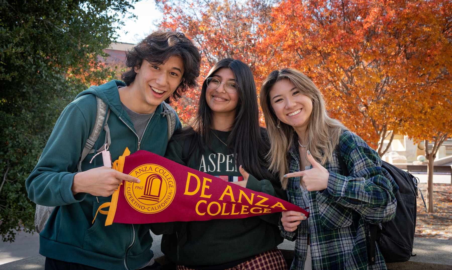 three students pointing to De Anza pennant and smiling